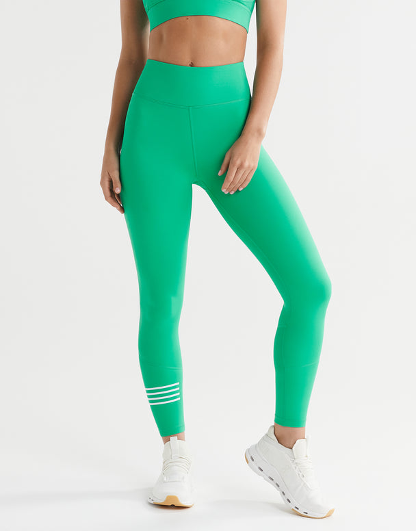 A woman wearing full-length leggings in electric green with a white printed stripe detail on the lower leg and a LILYBOD logo on the left ankle, paired with a sports bra and white sneakers.