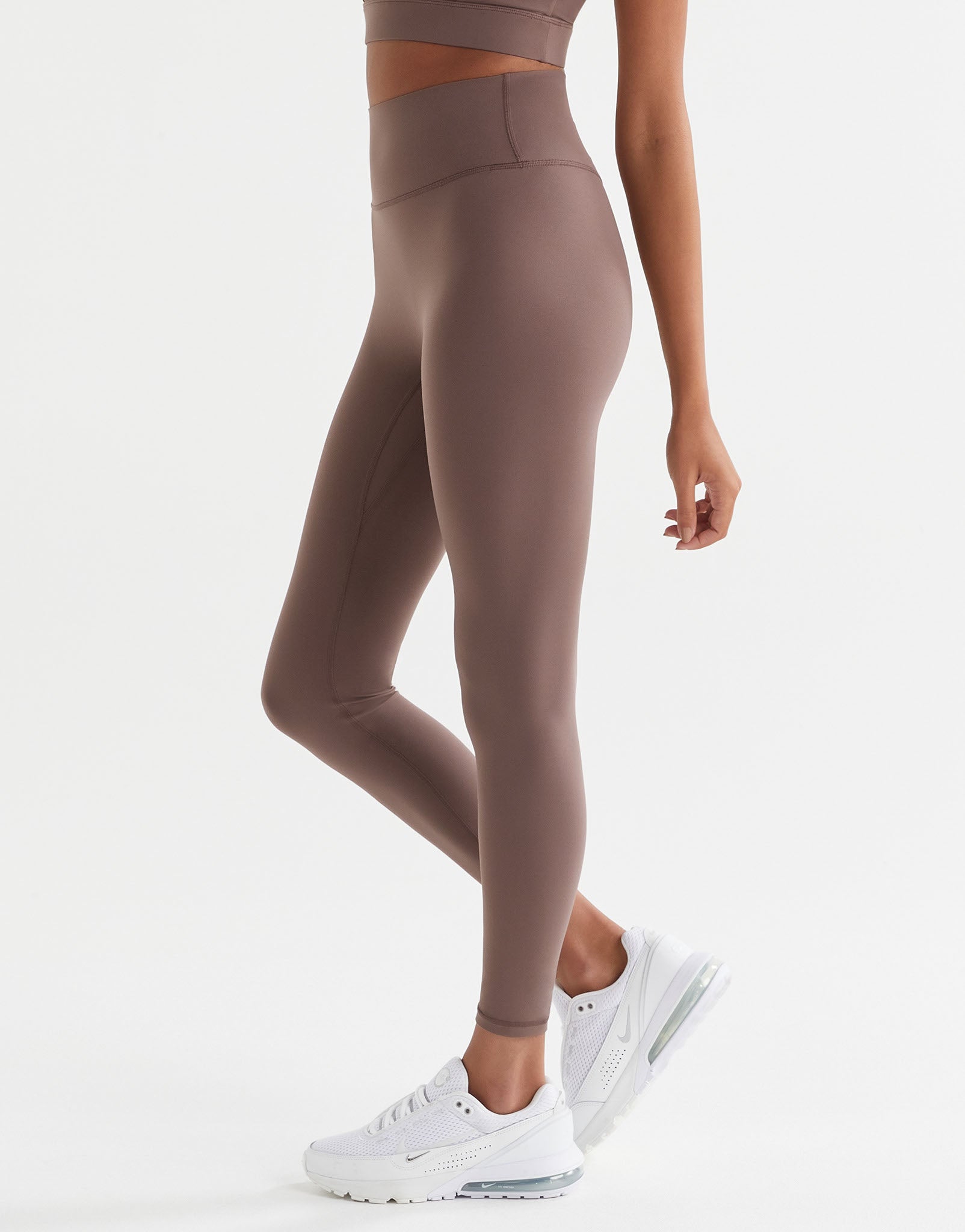 A woman standing wearing brown tinted, high-waisted leggings and a matching sports bra. The leggings have a minimal seaming design and are made from a fabric blend of polyamide and lycra.
