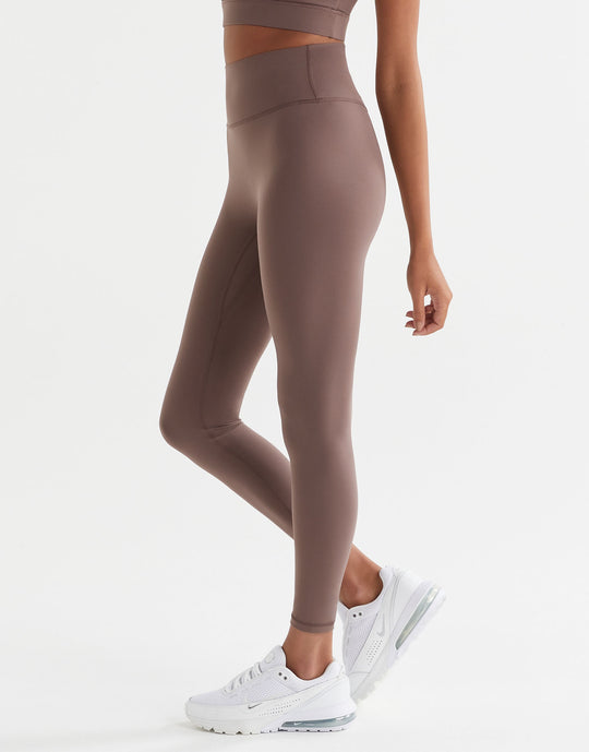 A woman standing wearing brown tinted, high-waisted leggings and a matching sports bra. The leggings have a minimal seaming design and are made from a fabric blend of polyamide and lycra.