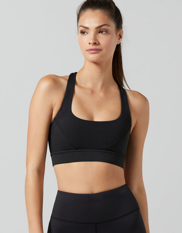 A woman modeling a black sports bra and matching high-waisted leggings against a solid light gray studio background.