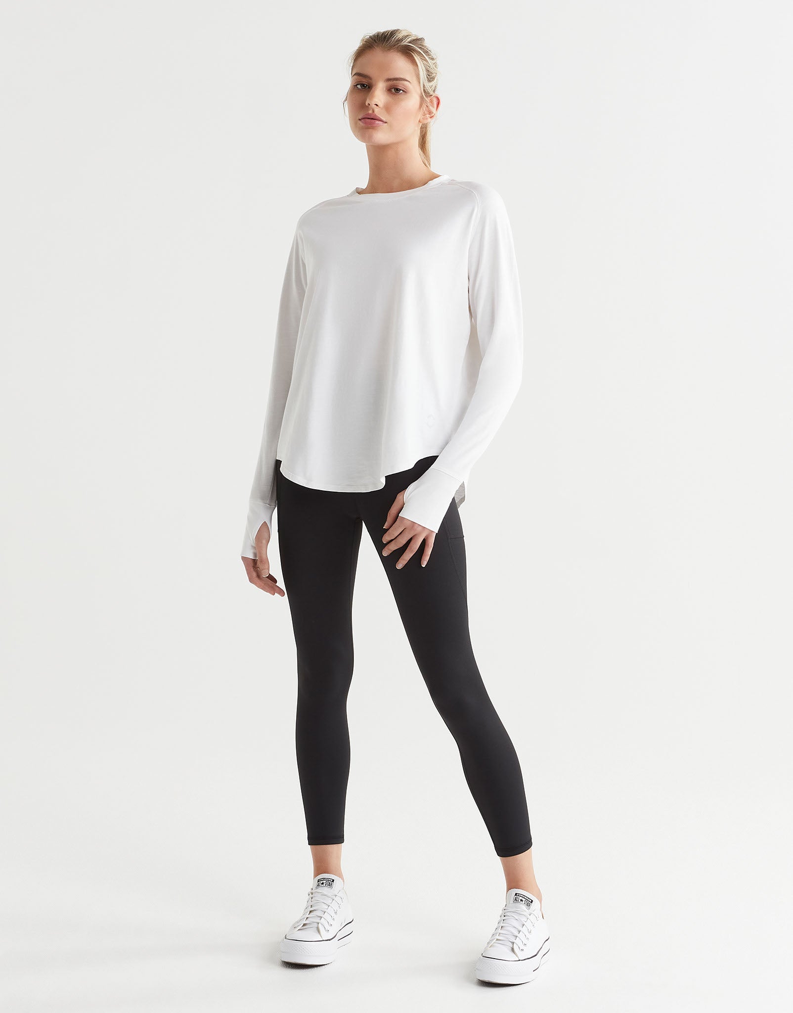 A female model wearing a long-sleeved white t-shirt with a round neckline, paired with black leggings and runners.