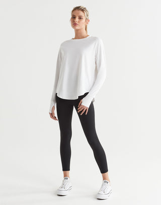 A female model wearing a long-sleeved white t-shirt with a round neckline, paired with black leggings and runners.