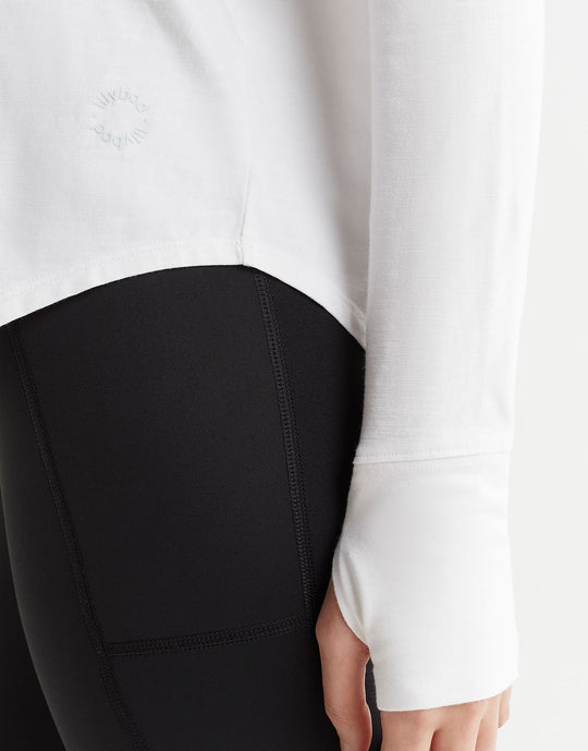 A female model wearing a long-sleeved white t-shirt with a round neckline and thumbholes, paired with black pants.
