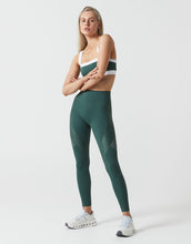 Woman wearing green leggings and a white sports bra with black straps on a light gray background