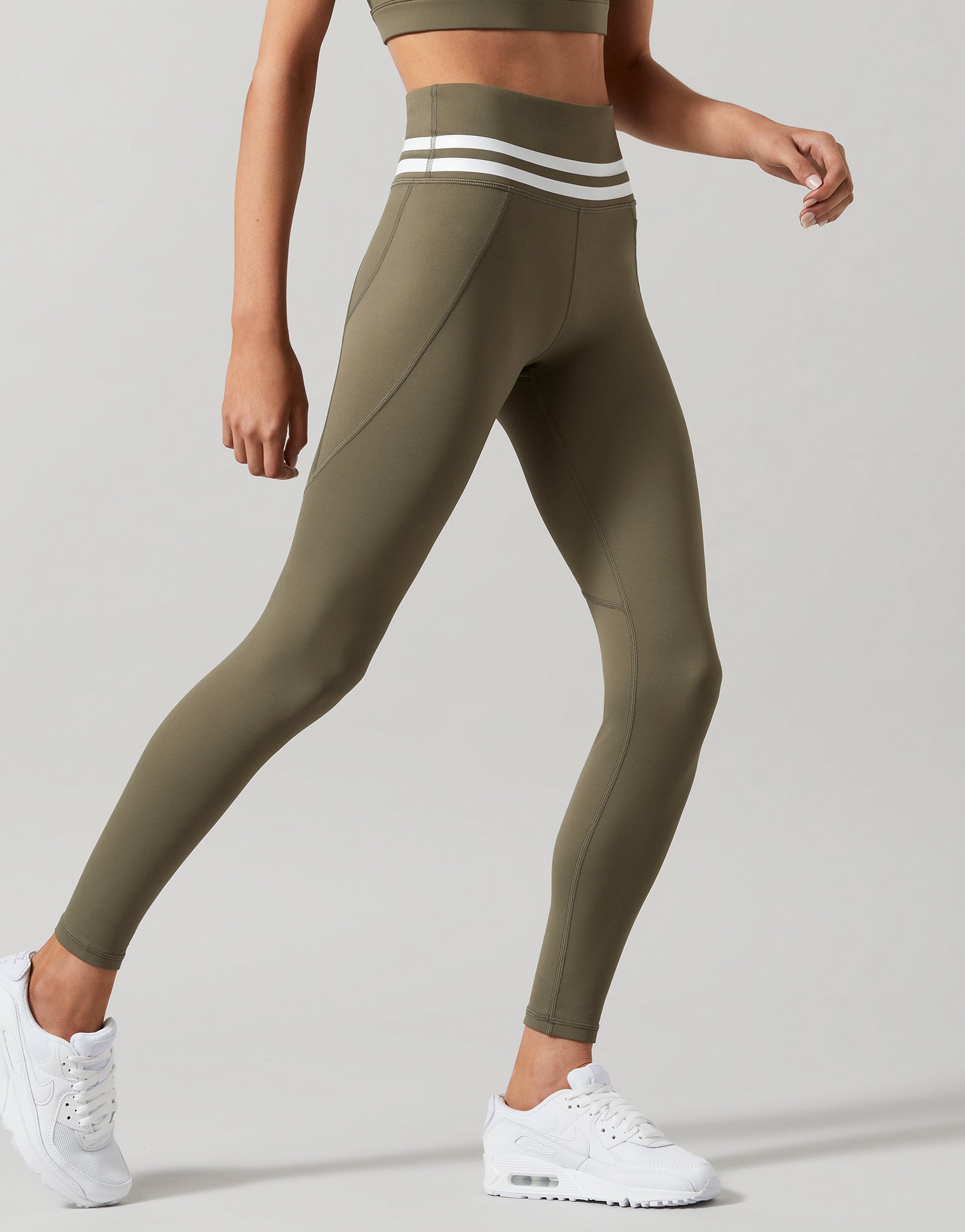 A pair of khaki green Lilybod Dakota leggings with a sports bra and white sneakers. Side view.