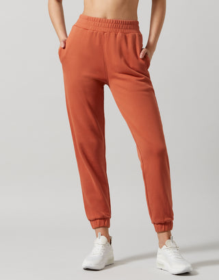 A pair of copper wash slim fit sweatpants with elastic waistband and cuffs, and a matching hoodie. The model is standing, wearing the sweatpants and white shoes.