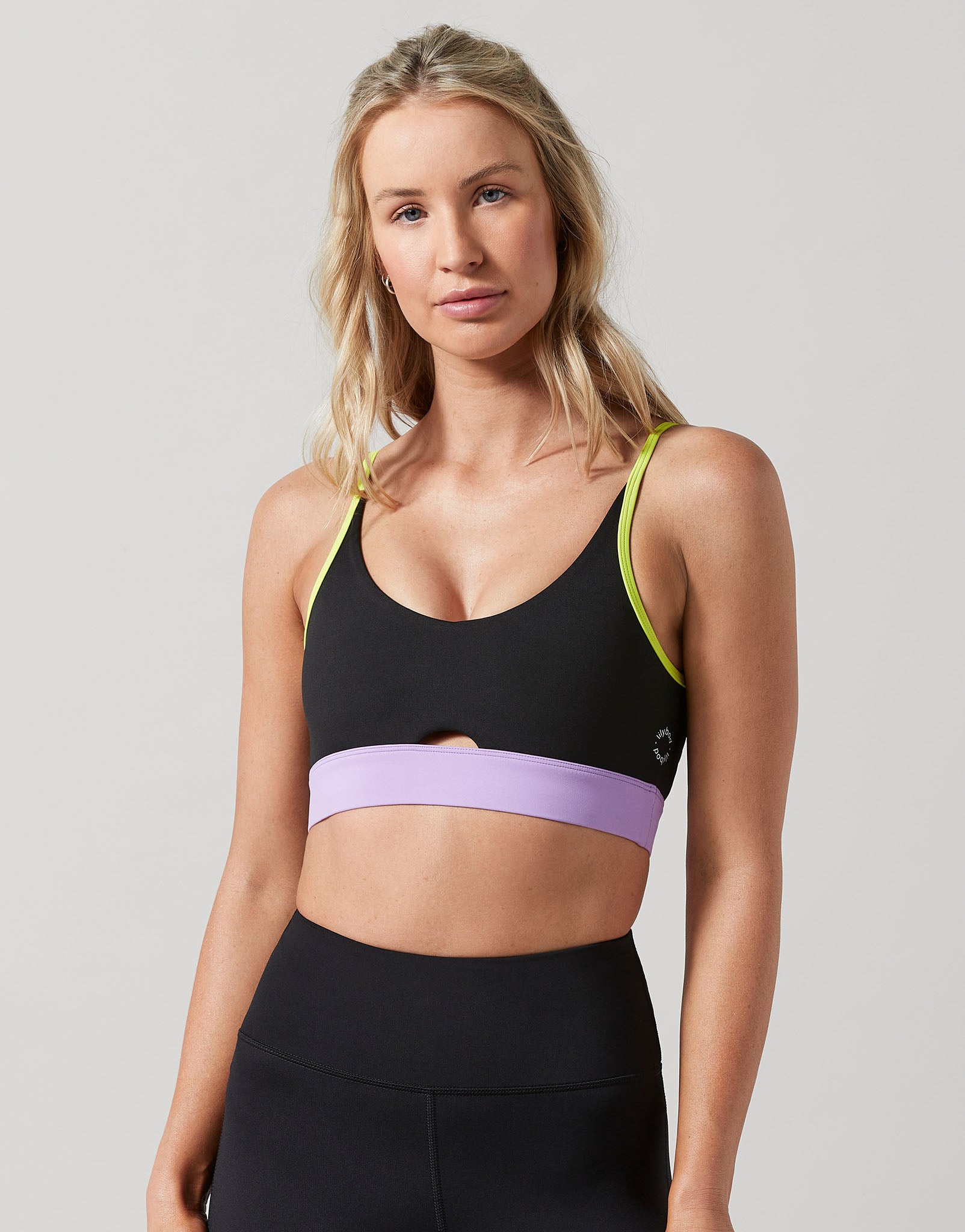 A woman modeling a black sports bra with a wide lavender band across the underbust and neon yellow-green trim along the straps, paired with black athletic leggings, against a light gray studio background.