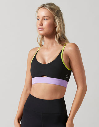 A woman modeling a black sports bra with a wide lavender band across the underbust and neon yellow-green trim along the straps, paired with black athletic bottoms, against a light gray studio background.