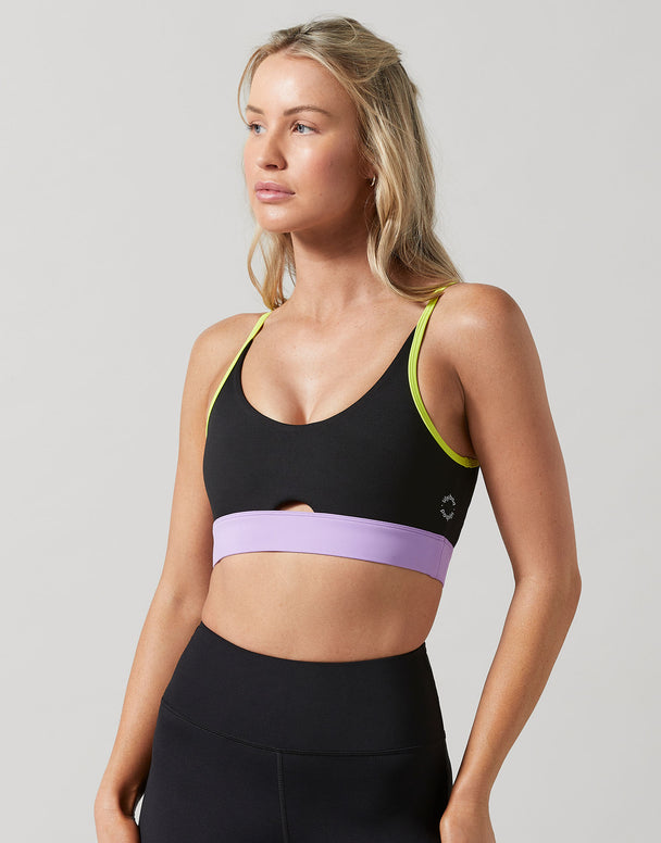 A woman modeling a black sports bra with a wide lavender band across the underbust and neon yellow-green trim along the straps, paired with black athletic bottoms, against a light gray studio background.