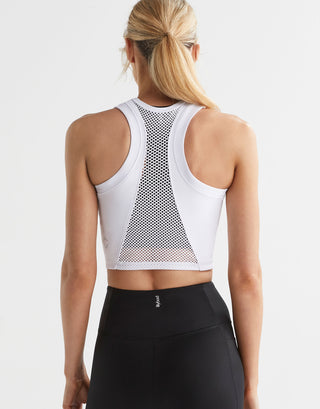 A female model wearing a white Lilybod Frankie Mesh Tank with a cropped fit and breathable mesh panels, featuring a racerback design and black leggings.