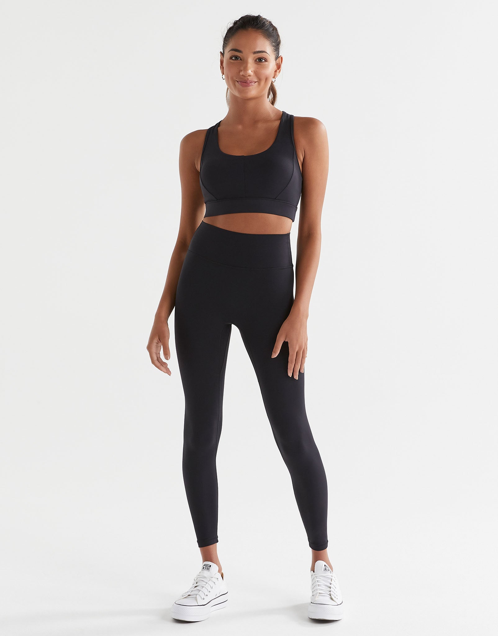 A woman wearing a black Lilybod Frankie Cloud-Core Sports Bra with wide straps and removable padded cups, leggings and white runners.