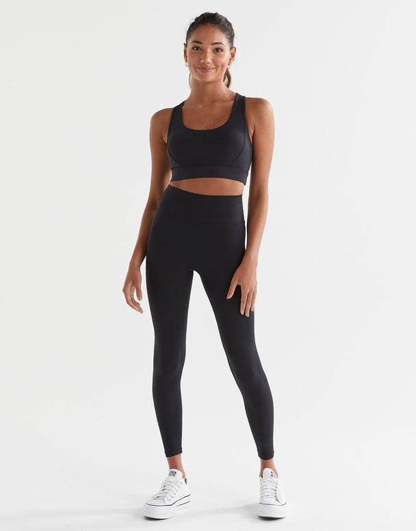 A woman wearing a black Lilybod Frankie Cloud-Core Sports Bra with wide straps and removable padded cups, leggings and white runners.