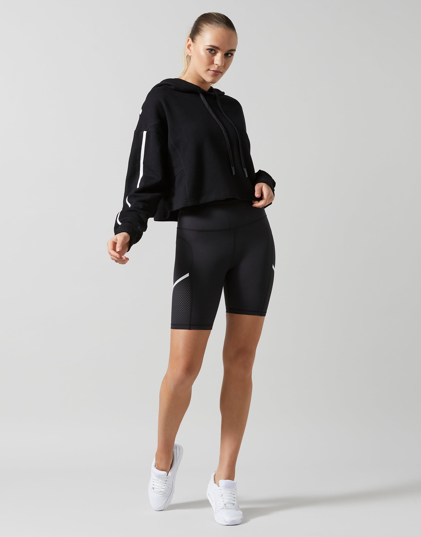 A female model wearing a smoke black oversized hoodie sweater with white printed sleeve hits and bike shorts with white sneakers.