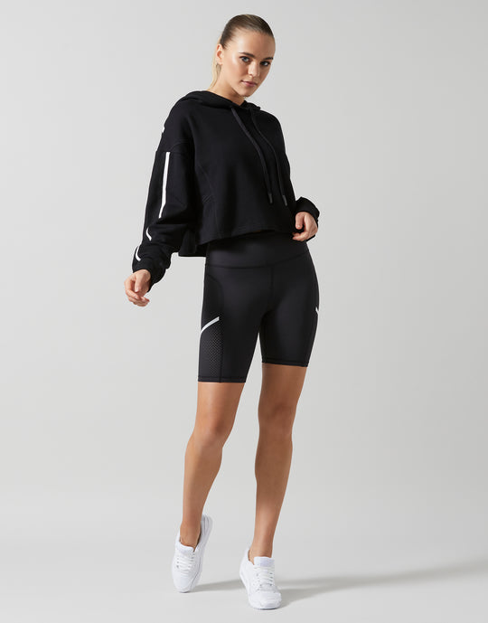 A female model wearing a smoke black oversized hoodie sweater with white printed sleeve hits and bike shorts with white sneakers.