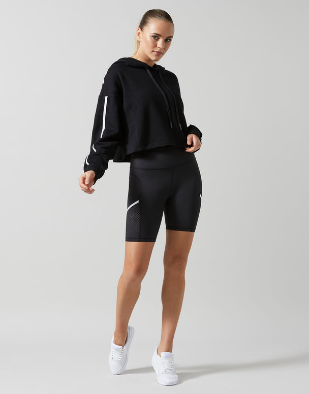 A female model wearing a smoke black oversized hoodie sweater with white printed sleeve hits and bike shorts with white sneakers.
