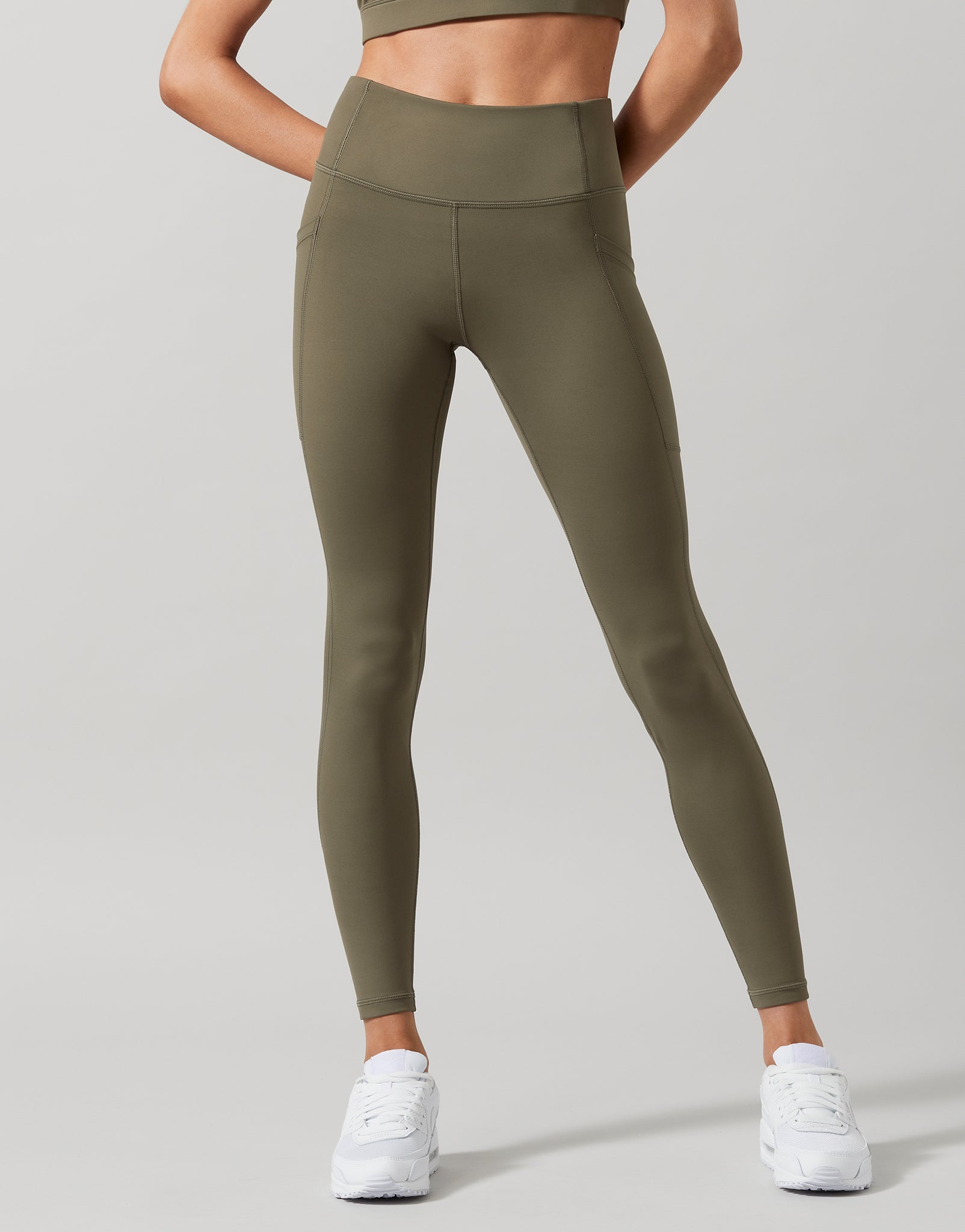 A model wearing high-waist full length khaki leggings with a Lilybod signature sculpting fabric, standing with hands in pockets.