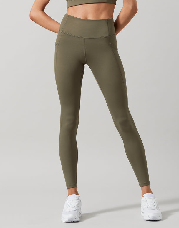 A model wearing high-waist full length khaki leggings with a Lilybod signature sculpting fabric, standing with hands in pockets.