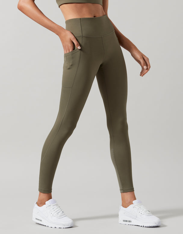 A woman wearing high-waist full length khaki leggings with a Lilybod signature sculpting fabric, standing with hands in pockets.