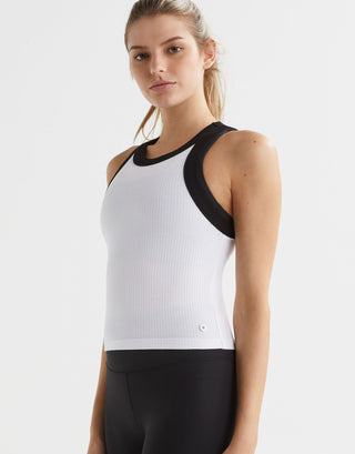 A female model wearing a white and black layered tank top with a slim-fit silhouette and contrast wide bind arm finish. Side view.
