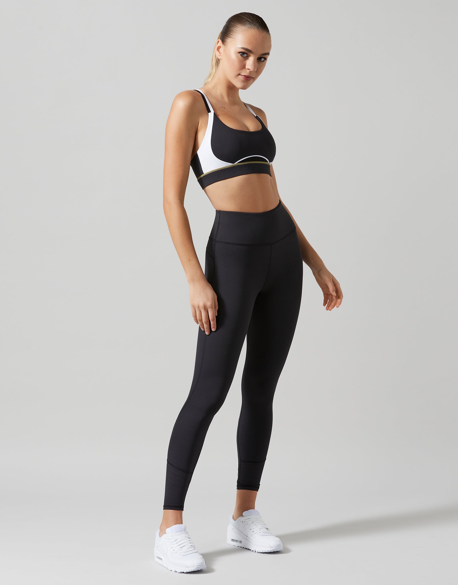 A woman modeling a black sports bra and matching high-waisted black leggings with white sneakers, standing against a light gray studio background.