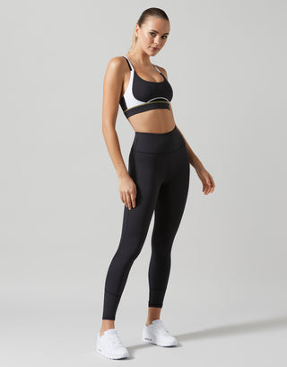 A woman modeling a black sports bra and matching high-waisted black leggings with white sneakers, standing against a light gray studio background.