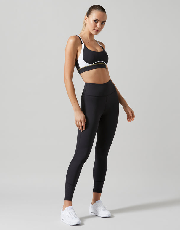 A woman modeling a black sports bra and matching high-waisted black leggings with white sneakers, standing against a light gray studio background.