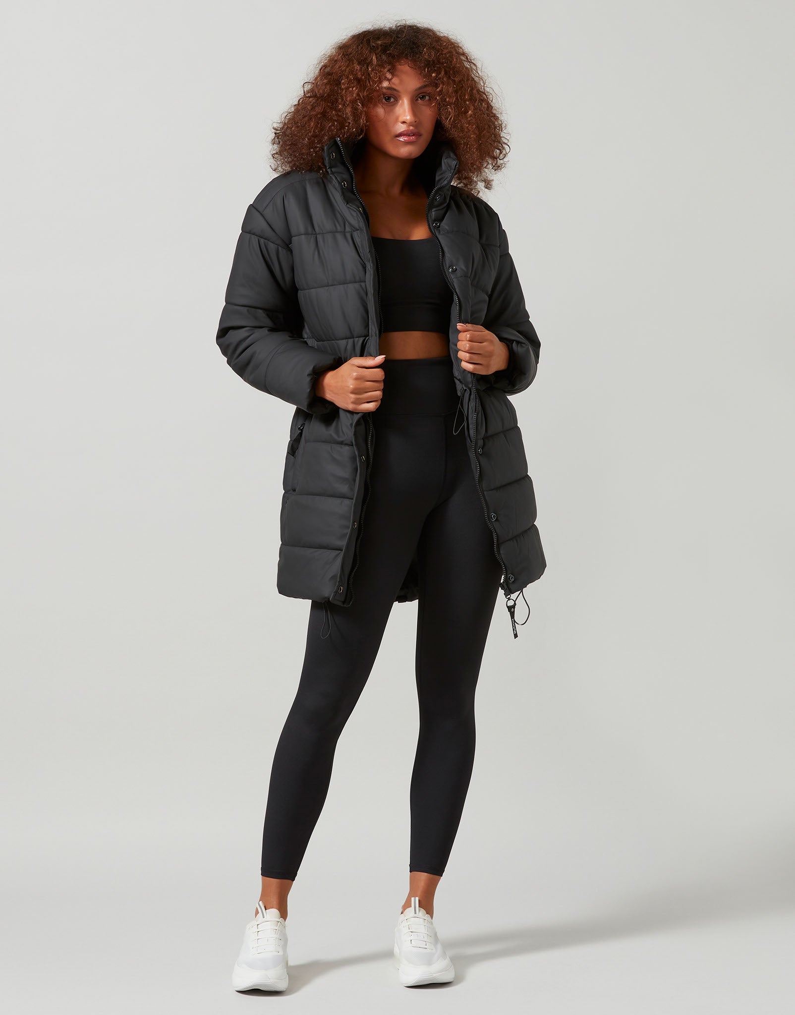 Woman wearing a black puffer jacket, black top, and leggings on a light gray background