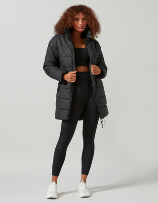 Woman wearing a black puffer jacket, black top, and leggings on a light gray background