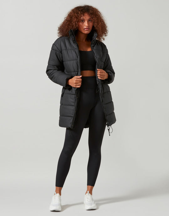 Woman wearing a black puffer jacket, black top, and leggings on a light gray background