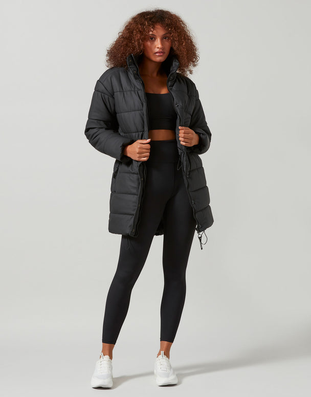 Woman wearing a black puffer jacket, black top, and leggings on a light gray background