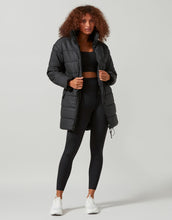 Woman wearing a black puffer jacket, black top, and leggings on a light gray background
