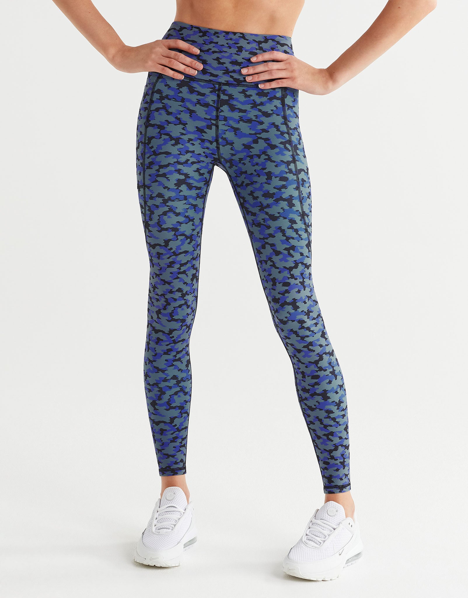 A woman wearing blue printed leggings with white sneakers on a white background.