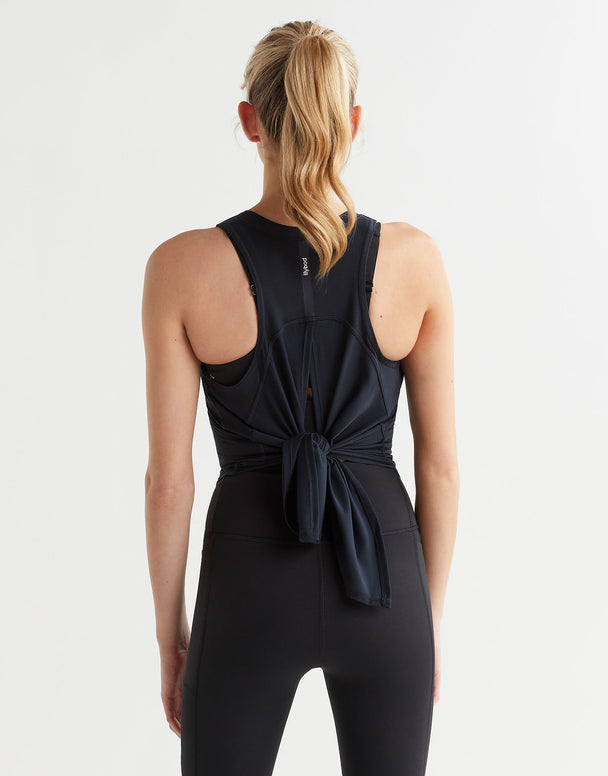 A woman viewed from the back wearing a black athletic jumpsuit with a knotted back detail, set against a plain light gray studio background.