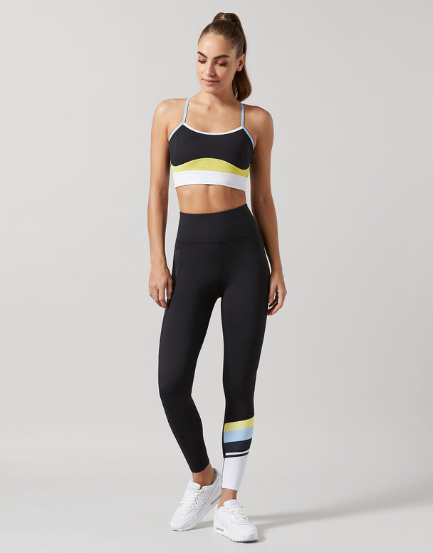 A woman modeling a matching black activewear set consisting of a sports bra and high-waisted leggings, featuring color-blocked details in white, yellow, and blue, paired with white sneakers on a light gray studio background.