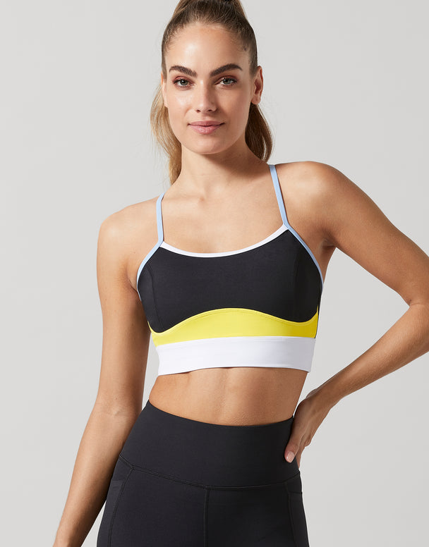 A woman modeling a black sports bra with a wavy colorblock pattern in yellow, white, and light blue, paired with black athletic leggings, against a solid light gray background.