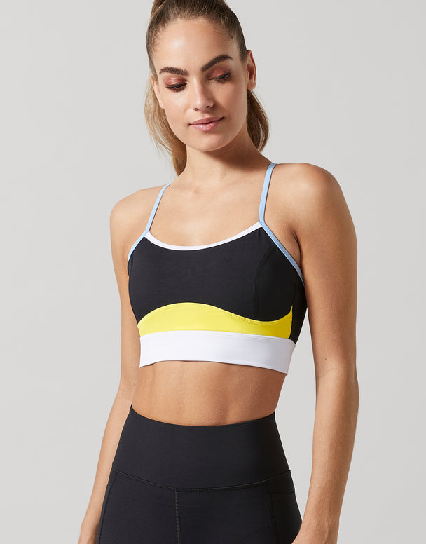A woman modeling a black sports bra with a yellow and white wavy stripe design across the bust, paired with black athletic leggings, against a light gray studio background.