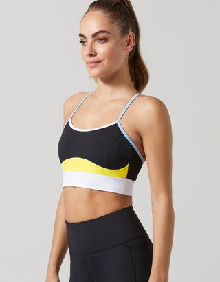 A woman modeling a black sports bra with a yellow and white wavy stripe design and light blue straps, paired with black athletic leggings, against a solid gray background.