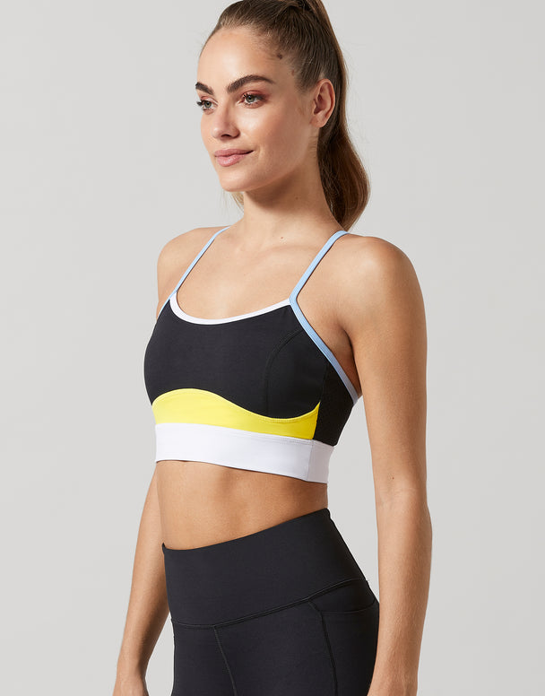 A woman modeling a black sports bra with a yellow and white wavy stripe design and light blue straps, paired with black athletic leggings, against a solid gray background.