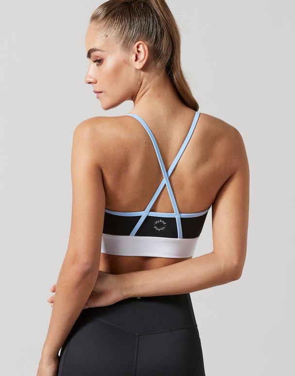 A woman shown from the back wearing a black sports bra with light blue and white trim, paired with black leggings, against a solid light gray background.