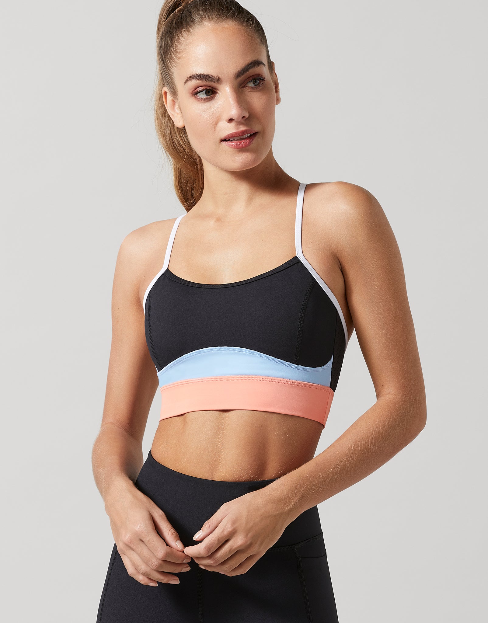 A woman modeling a color-blocked sports bra with a black upper section, a light blue middle band, and a peach-pink lower band, paired with black leggings against a solid gray background.