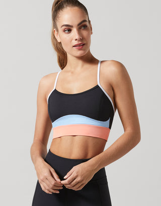 A woman modeling a color-blocked sports bra with a black upper section, a light blue middle band, and a peach-pink lower band, paired with black leggings against a solid gray background.