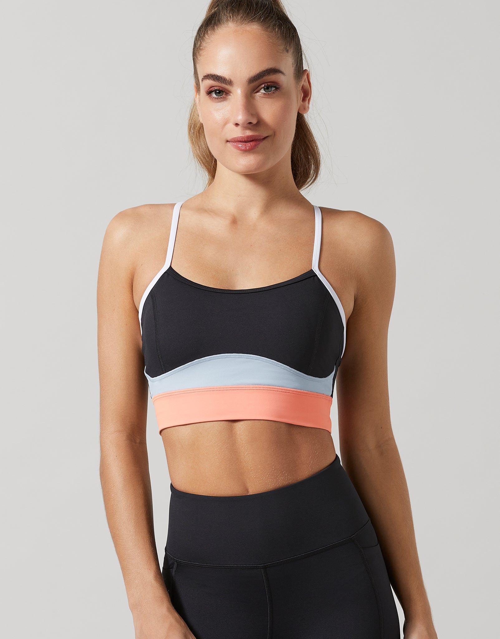 A woman modeling a color-blocked sports bra with black, light blue, and coral pink bands, paired with black athletic leggings, against a solid gray background.