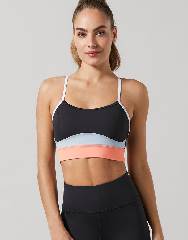 A woman modeling a color-blocked sports bra with black, light blue, and coral pink bands, paired with black athletic leggings, against a solid gray background.