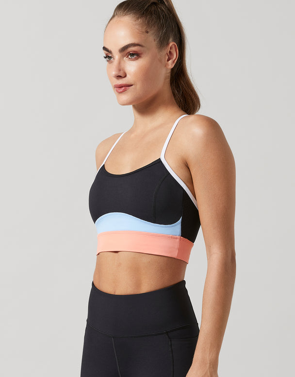 A woman wearing a black sports bra with a color-blocked design featuring white, light blue, and peach-pink horizontal bands, paired with black leggings, against a solid light gray background.