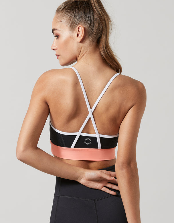 A woman shown from the back wearing a sports bra with white crisscross straps, a black upper band, and a wide metallic pink band at the bottom, paired with black leggings against a light gray studio background.