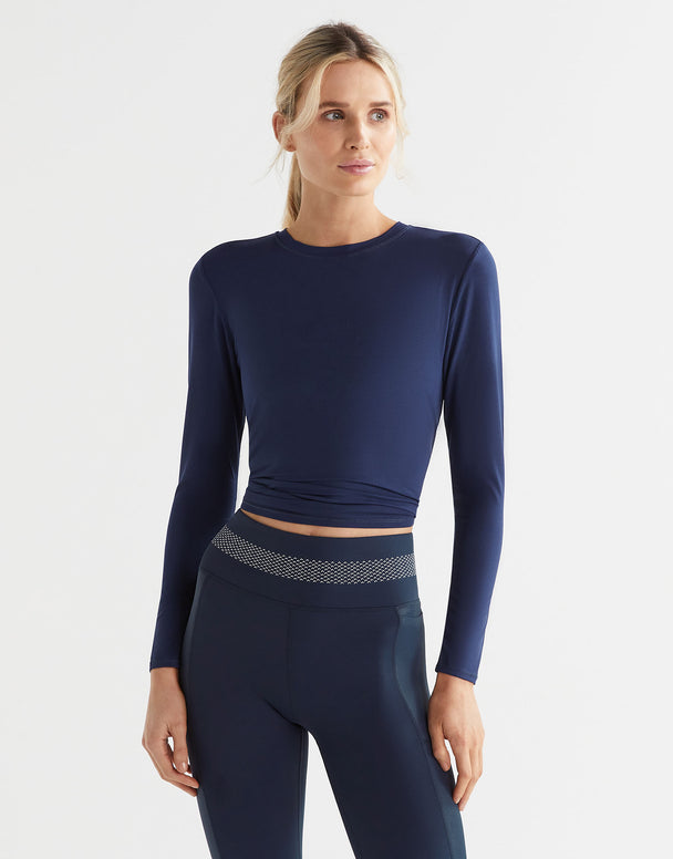 A woman wearing a navy long-sleeved workout top with a tie-up detail at the waist, paired with matching navy leggings.