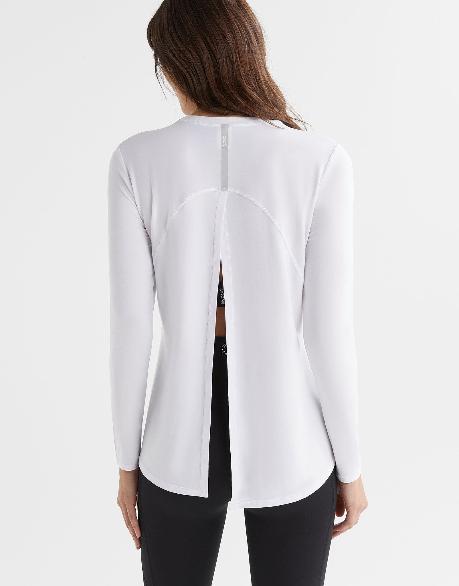 A woman wearing a bright white long sleeved activewear top with a relaxed fit. The top has a crew neckline and a split back that can be tied up.