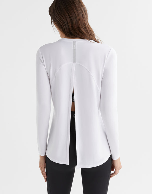 A woman wearing a bright white long sleeved activewear top with a relaxed fit. The top has a crew neckline and a split back that can be tied up.