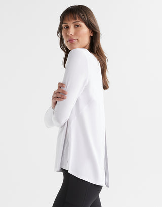 A model wearing a bright white long sleeved activewear top with a relaxed fit. The top has a crew neckline and a split back that can be tied or left open.