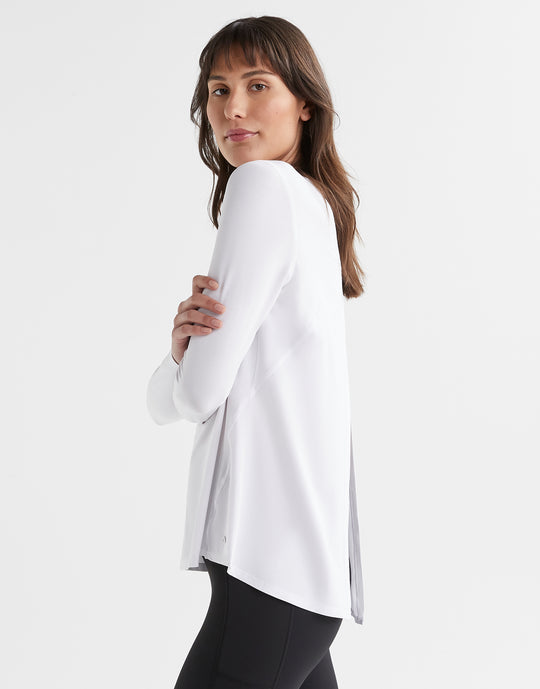A model wearing a bright white long sleeved activewear top with a relaxed fit. The top has a crew neckline and a split back that can be tied or left open.